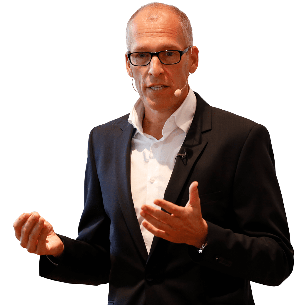 Ein Mann im Anzug und mit Brille spricht gestikulierend mit einem Headset-Mikrofon vor einem schlichten Hintergrund.